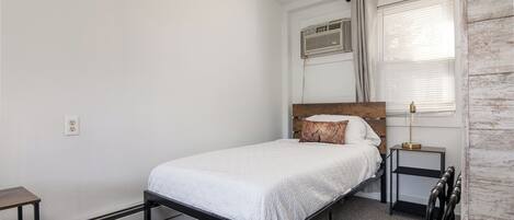A single bed with a white quilt, a bedside table, a nightstand with a lamp, a window with curtains, and a wall-mounted air conditioning unit.