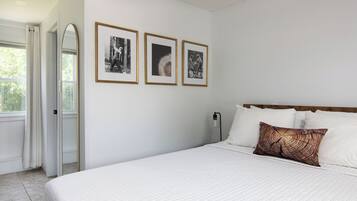 Quarto com cama, roupa de cama branca, cabeceira de madeira, mesa de cabeceira, luminária na parede e três quadros emoldurados.