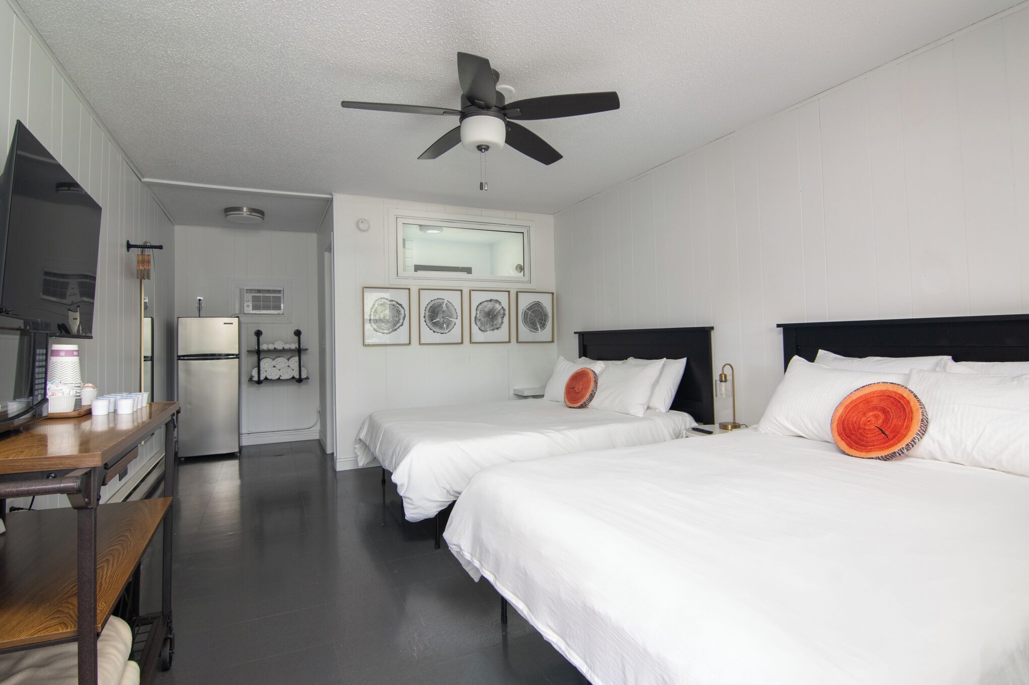 Habitación de hotel con dos camas, ventilador de techo, refrigerador, escritorio con silla y espejo.