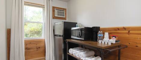A compact kitchenette with a refrigerator, microwave, and coffee maker, a wooden bench with towels, and a window with curtains.