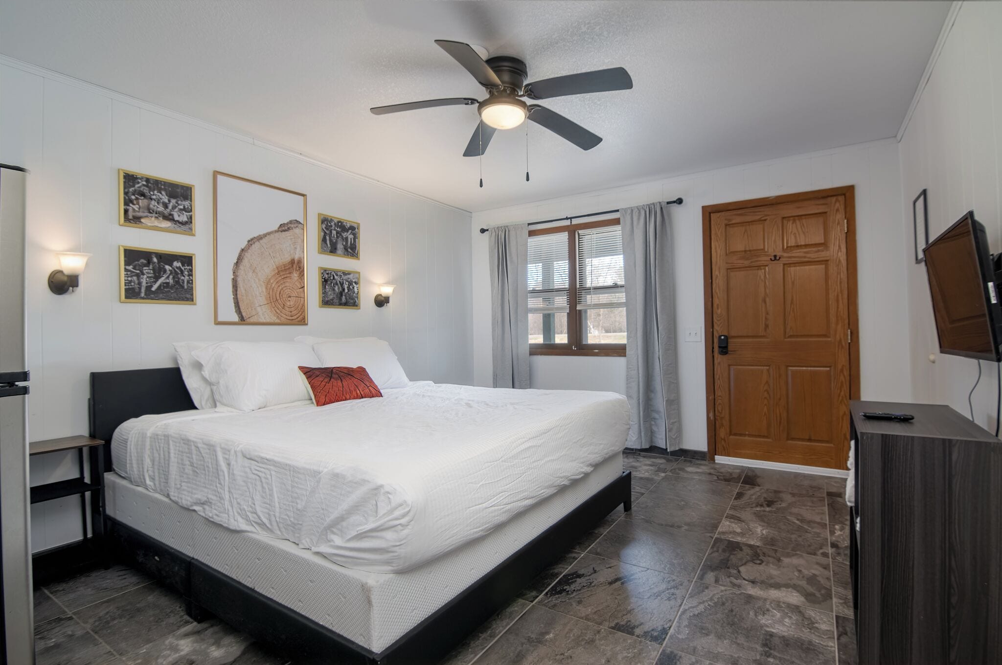 A bedroom with a bed, ceiling fan, artwork, window, door, and a TV stand.