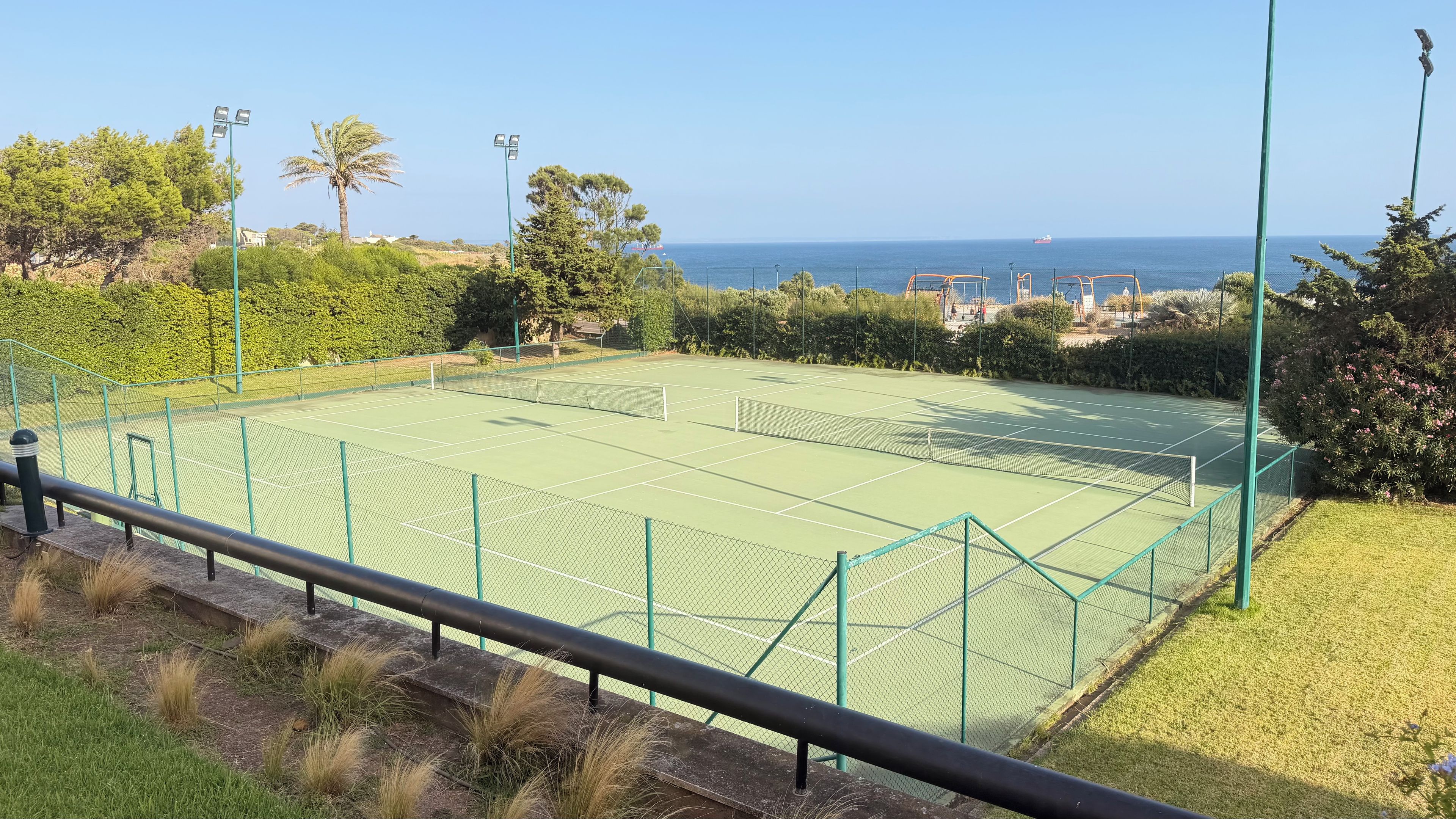 Ein Tennisplatz mit Netz, umzäunt und mit Blick auf ein Gewässer, in der Ferne ein Schiff.