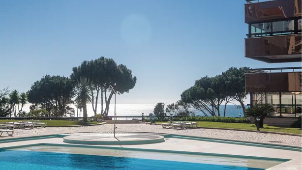 Ein Poolbereich mit Blick auf das Meer, umgeben von Bäumen und einem Gebäude mit großen Fenstern.
