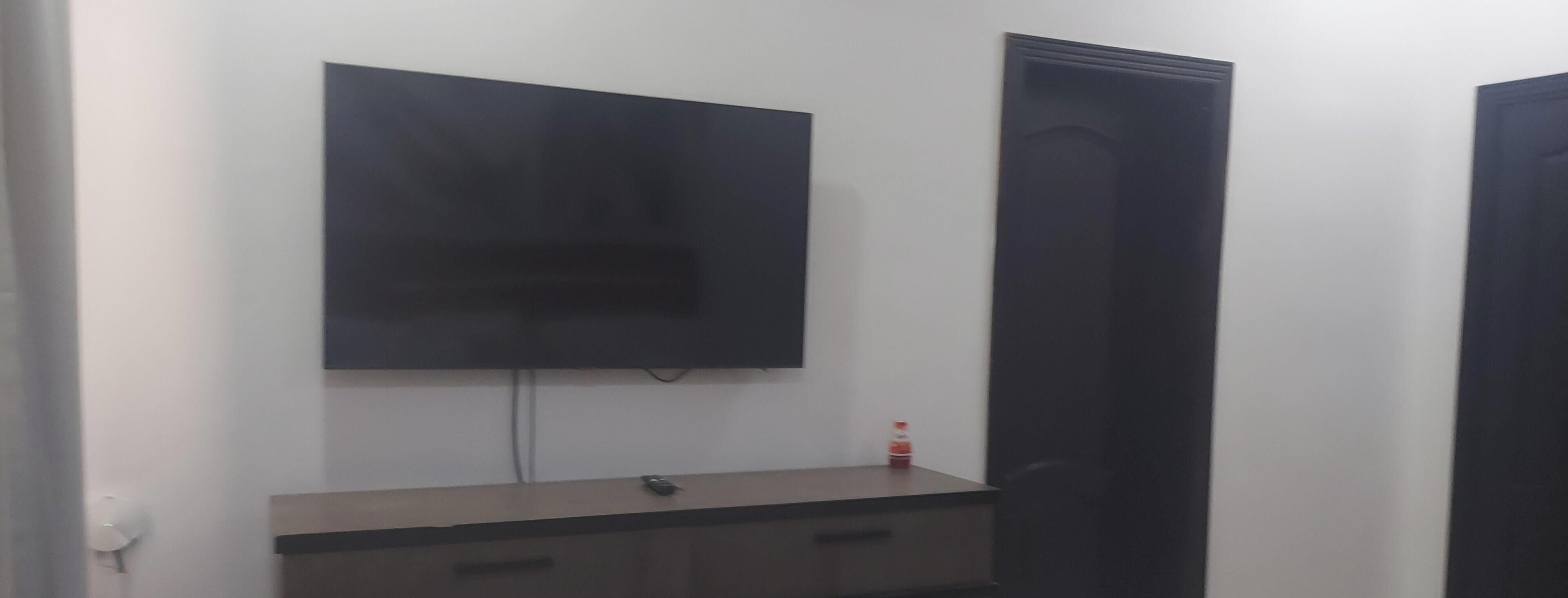 Ett rum med en platt-TV som är monterad på väggen, en byrå i trä med lådor, en dörr och en liten flaska som står på byrån.