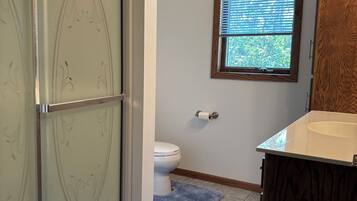 A bathroom with a glass shower enclosure, a toilet, a window with blinds, and a wooden vanity.