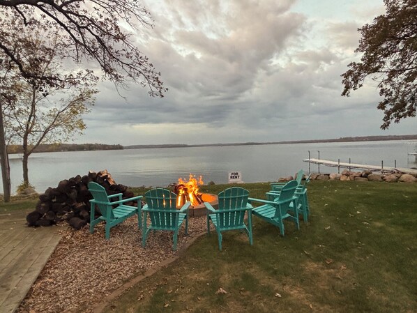 A lakeside seating area with a fire pit and chairs, a pile of firewood, and a 'For Rent' sign.