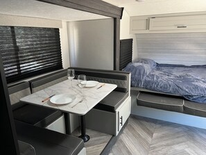 Quarto compacto com cama, mesa pequena com dois pratos e duas taças, e um banco.