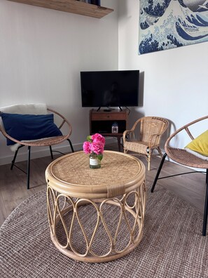 A room with a round wicker table, two wicker chairs, a television on a wooden stand, and a wall-mounted artwork.