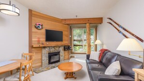 A cozy living room with a stone fireplace, a flat-screen TV mounted above it, a wooden dining table with chairs, a leather sofa with cushions, and a ski rack against the wall.