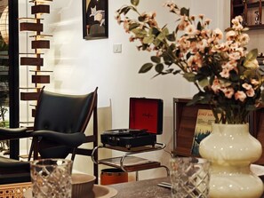 Un salon moderne avec un fauteuil en cuir noir, une table en verre, un vase de fleurs, un tableau encadré et une boîte rouge portant une signature.
