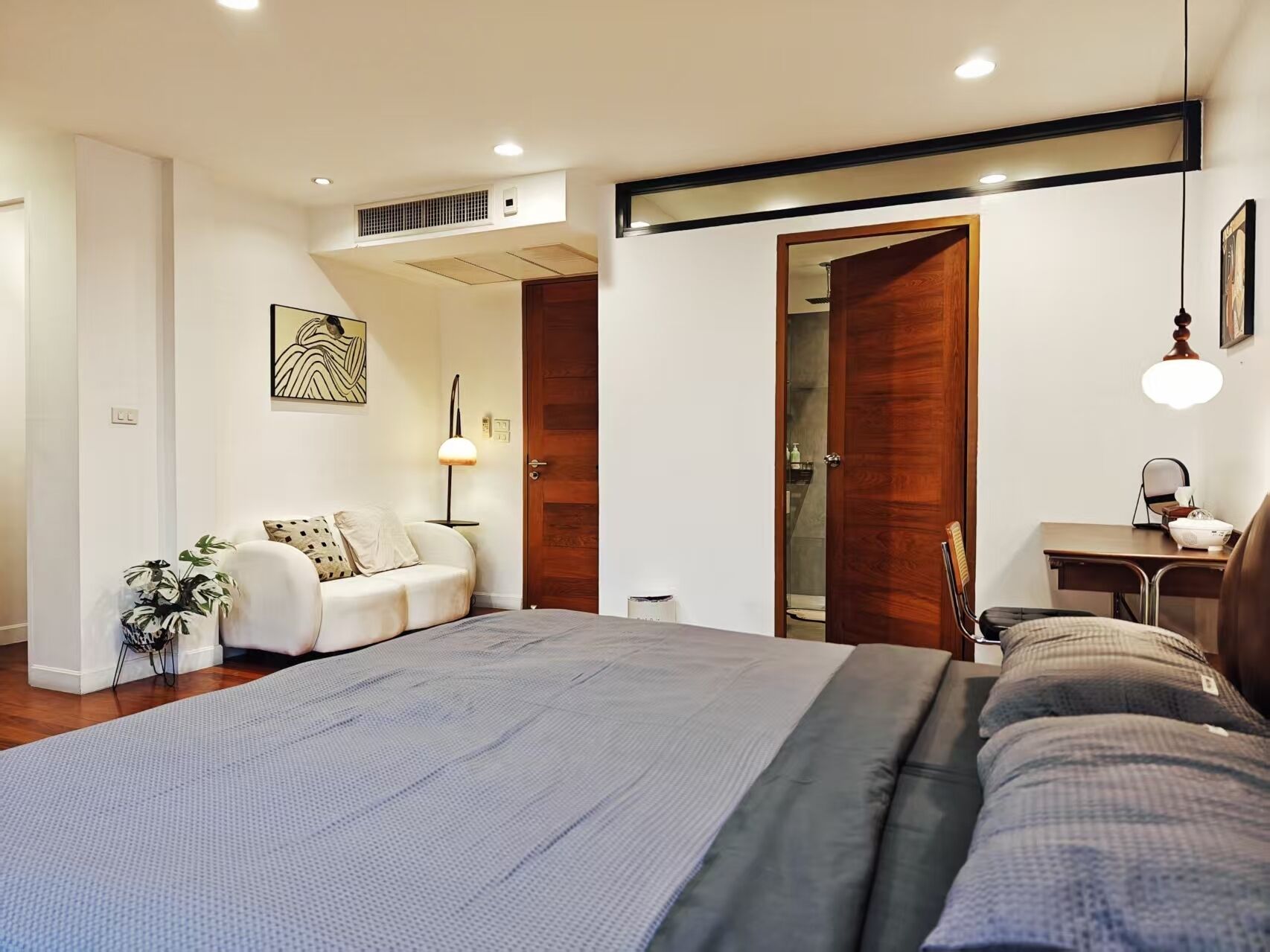 A modern bedroom with a large bed, a desk, and a chair. There is a plant and a painting on the wall.