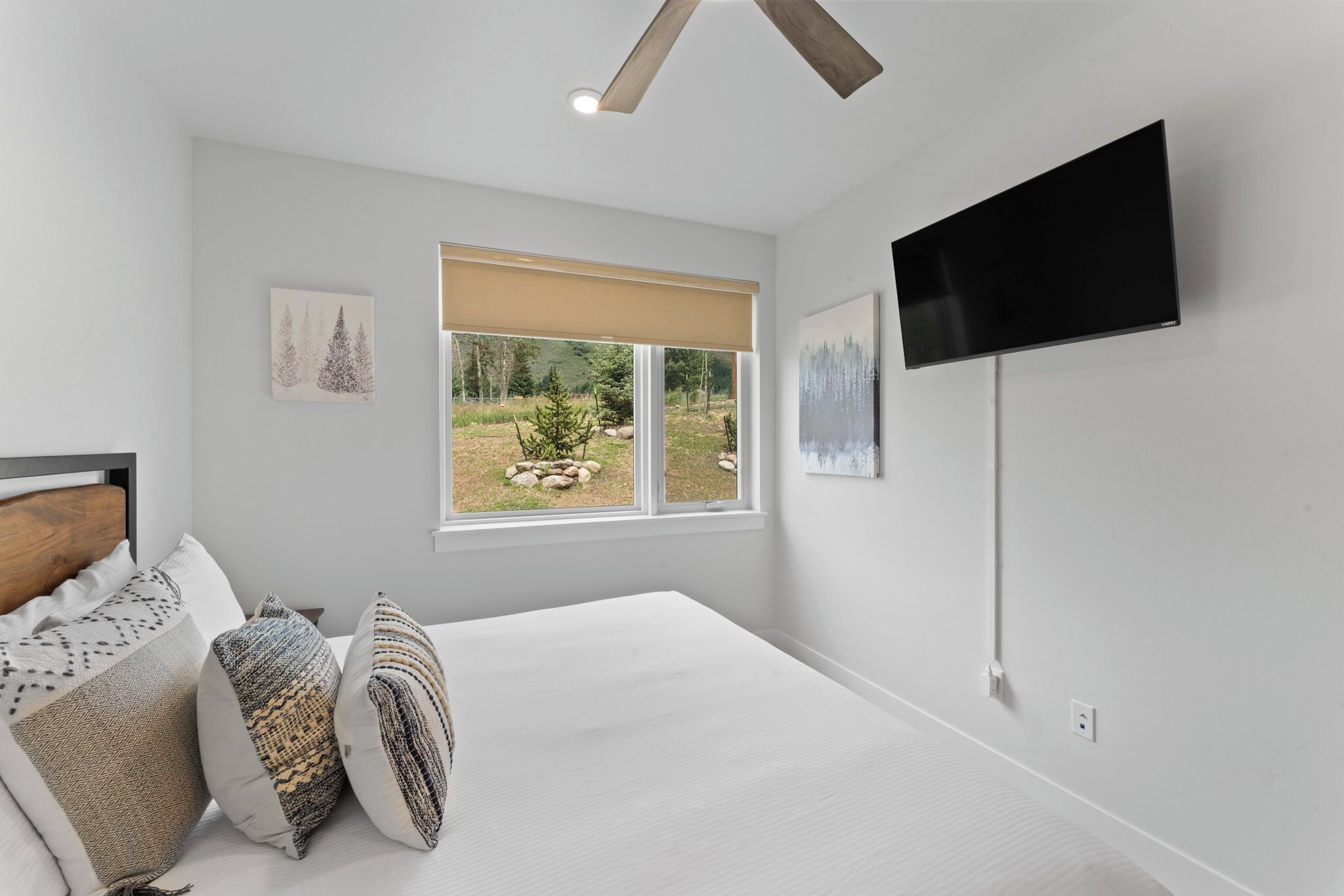Une chambre dotée d’un grand lit, d’un ventilateur de plafond, d’un téléviseur fixé au mur et d’une fenêtre donnant sur des arbres et un jardin.