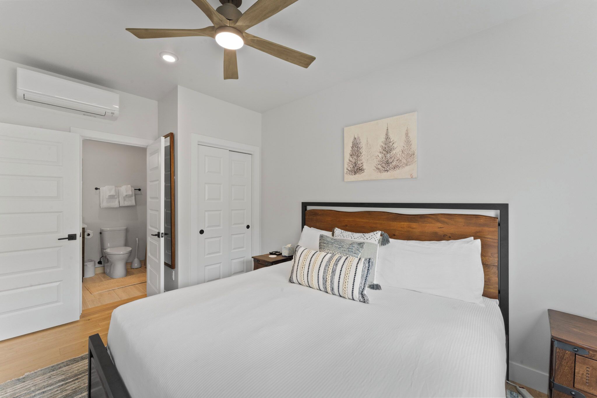 A bedroom with a bed, a wooden headboard, a ceiling fan, a white door, and a painting of trees.