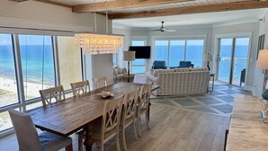 Amplia sala de estar con una mesa de comedor de madera y sillas, un sofá, y vista al océano a través de grandes ventanales.