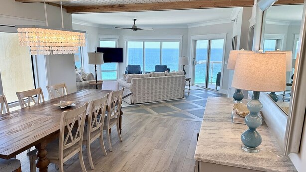 A spacious living area with a wooden dining table, chairs, a sofa, and a TV. The room has large windows offering a view of the ocean.