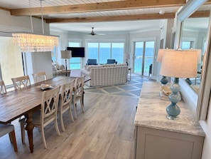 A spacious living area with a wooden dining table, chairs, a sofa, and a TV. The room has large windows offering a view of the ocean. - New Emerald Isle #1601 •Panoramic Views • Sleeps 8 (Pensacola Bach)