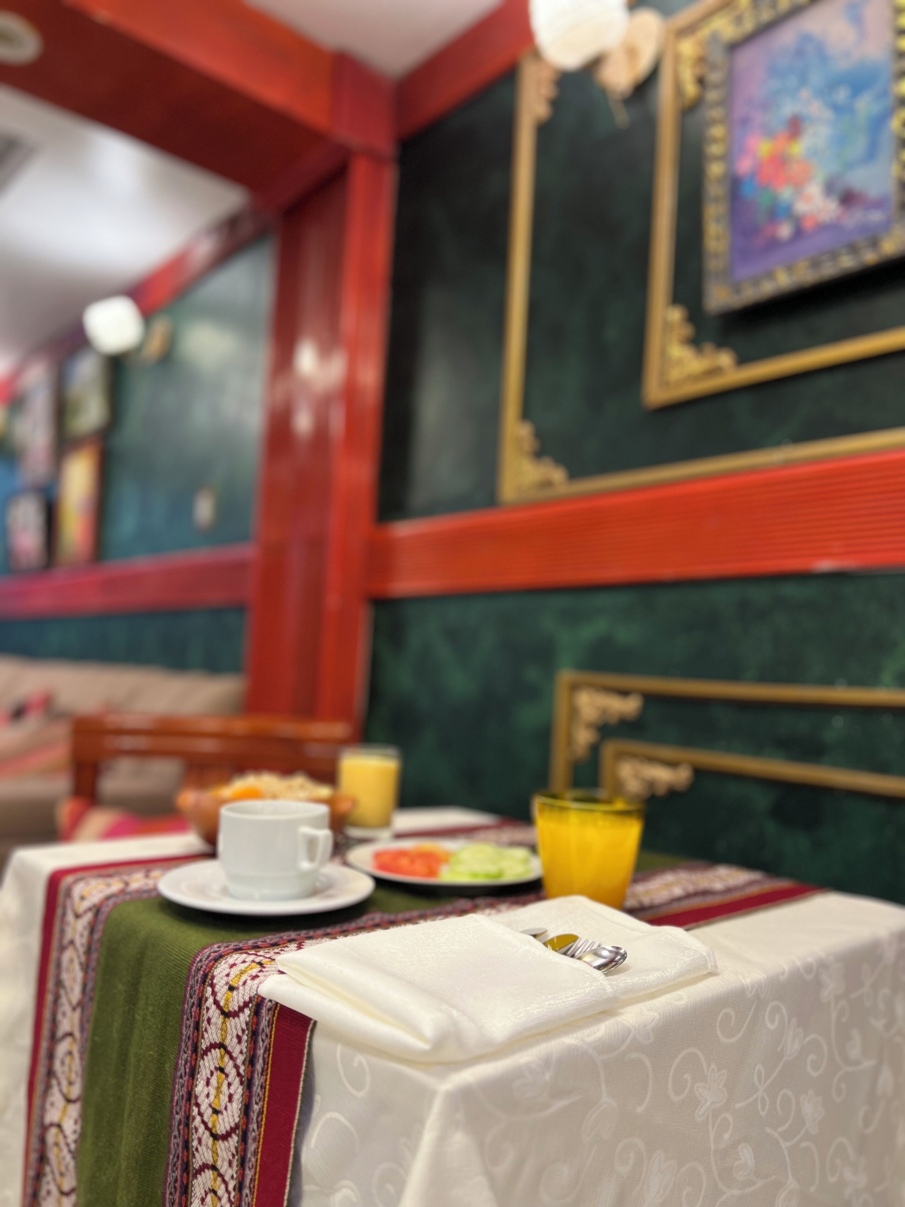 A dining table with a white tablecloth, a green patterned table runner, and a white napkin. There is a plate with food and a glass of orange juice. The room has red walls and a framed picture on the wall.