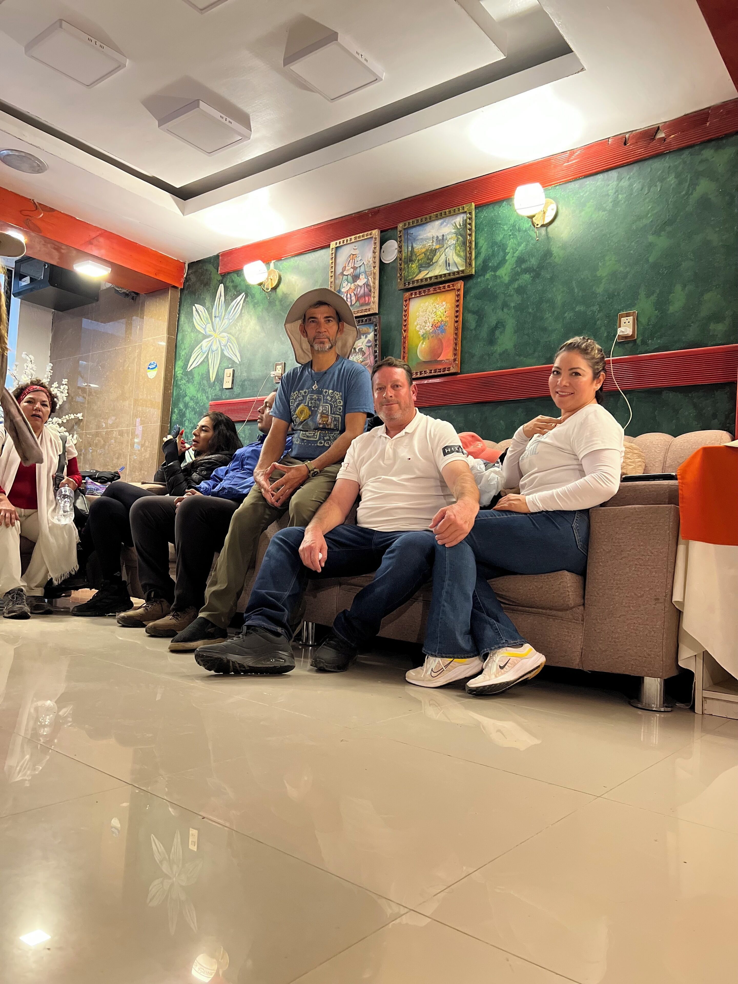 A group of people seated on a couch in a room with a green wall and red accents, a tiled floor, and ceiling lights.