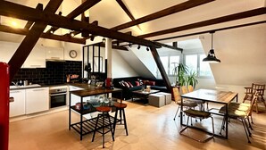 Un espacio habitable tipo loft con vigas de madera a la vista, una zona de cocina, una mesa de comedor con sillas y un área de estar con sofá y mesa de centro.