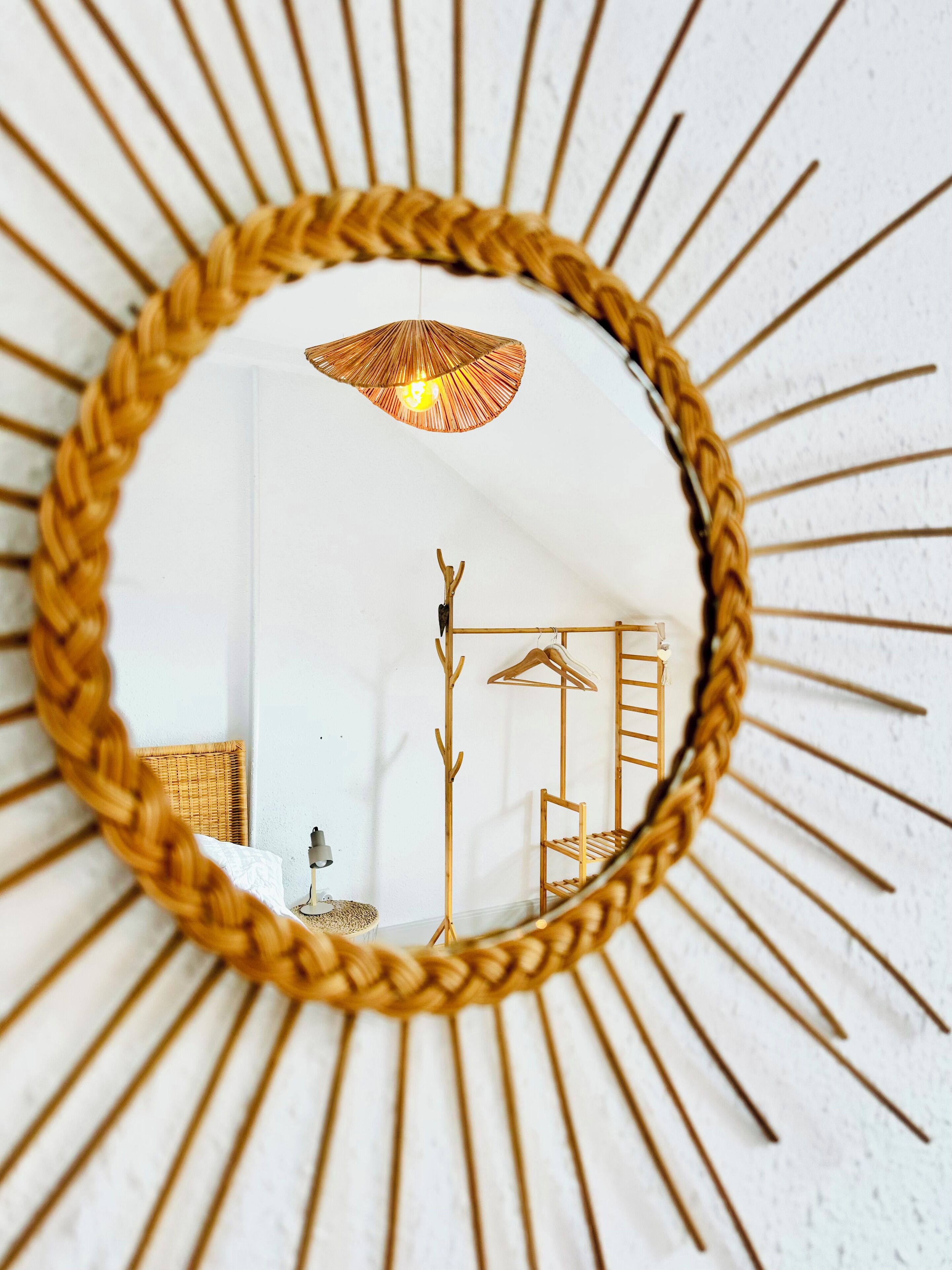 A sunburst mirror with a woven frame reflecting a room with a hanging light fixture, wooden coat rack, and a wicker chair.