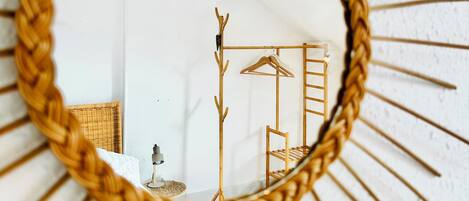 A sunburst mirror with a woven frame reflecting a room with a hanging light fixture, wooden coat rack, and a wicker chair.