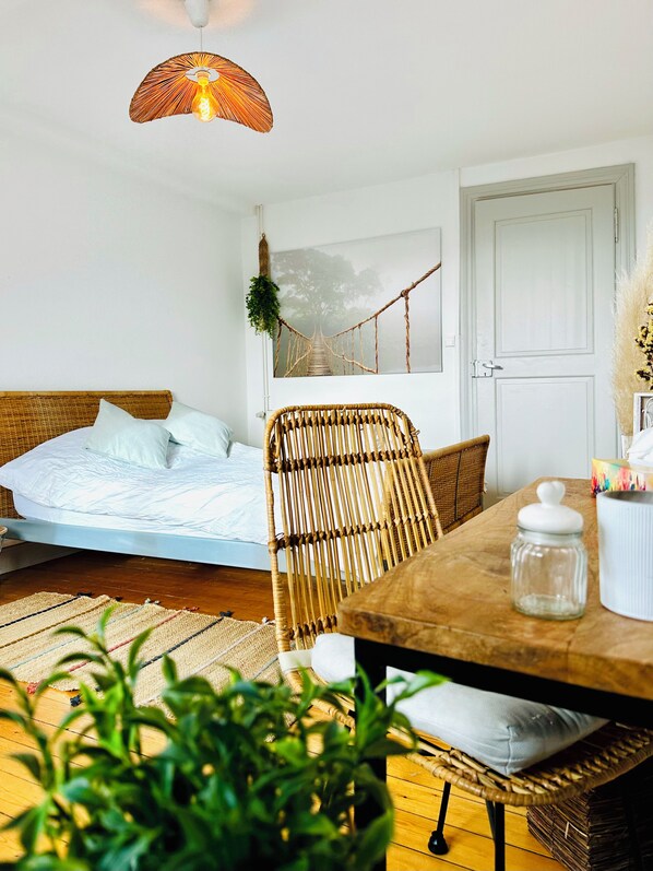 Ein Zimmer mit einem Bett, einem Esstisch, einem Korbstuhl, einer Pendelleuchte und einer Tür.