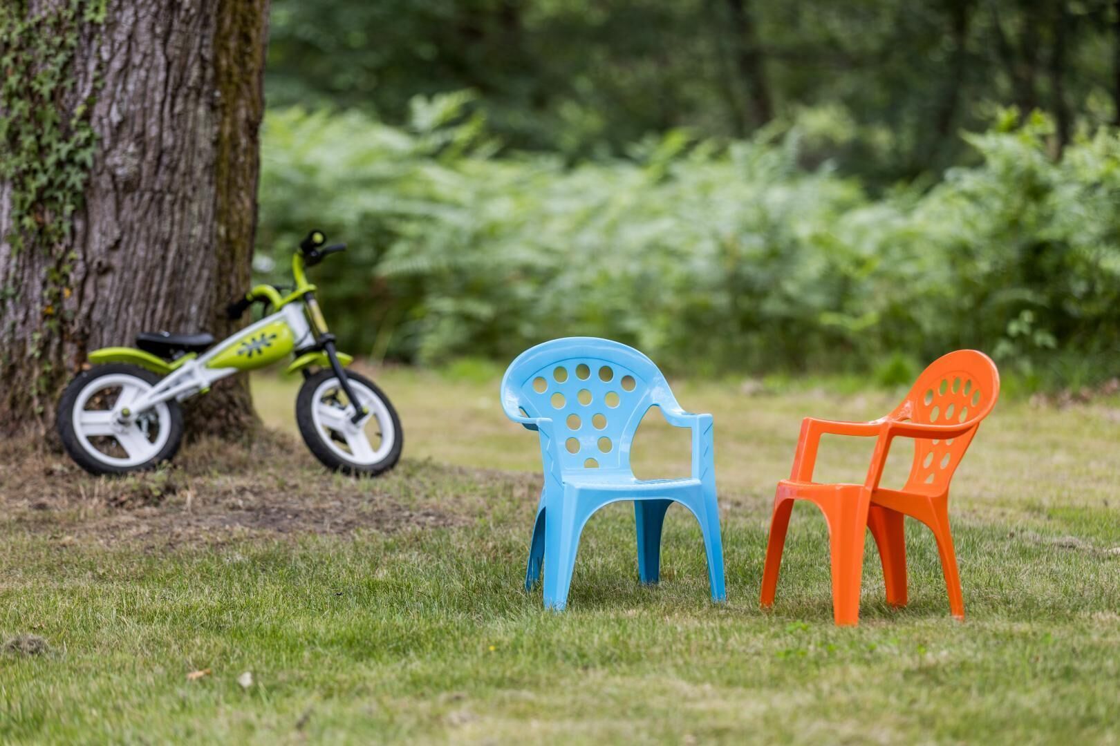 Deux chaises en plastique, une bleue et une orange, sont placées sur une pelouse. On aperçoit un arbre et un vélo vert en arrière-plan.