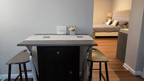 Una habitación compacta con un escritorio de madera oscura, dos taburetes, una pequeña mesa blanca con una caja de pañuelos y una cama con cabecera gris al fondo.