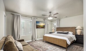 Ein Schlafzimmer mit Bett, Deckenventilator, Nachttisch, Lampe und einem Fenster mit Vorhängen.