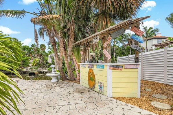 Un bar tiki con un letrero de madera, una fuente de piedra y plantas tropicales.