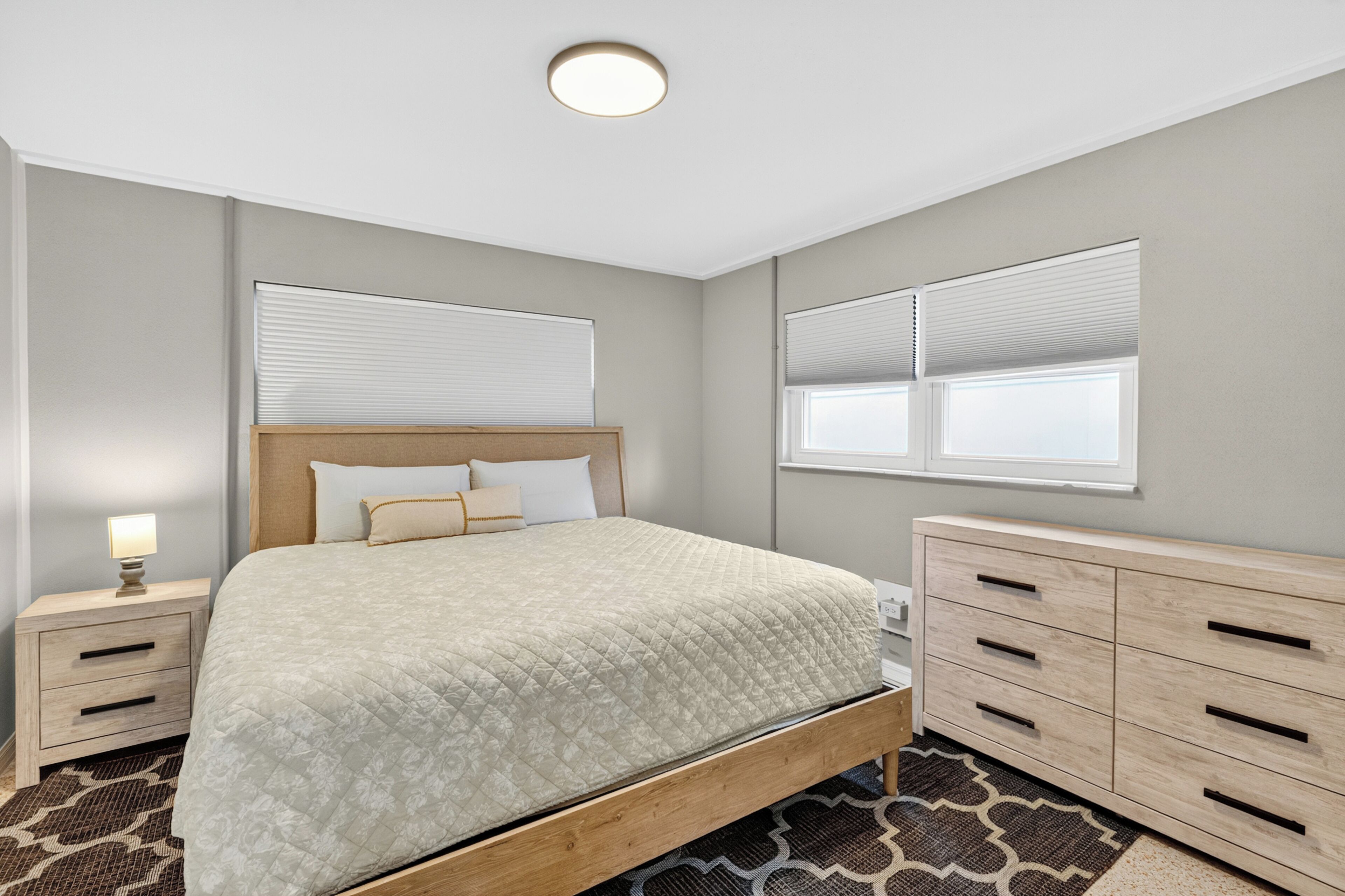 Un dormitorio con cama, mesitas de noche, cómoda, ventana con persianas e iluminación en el techo.