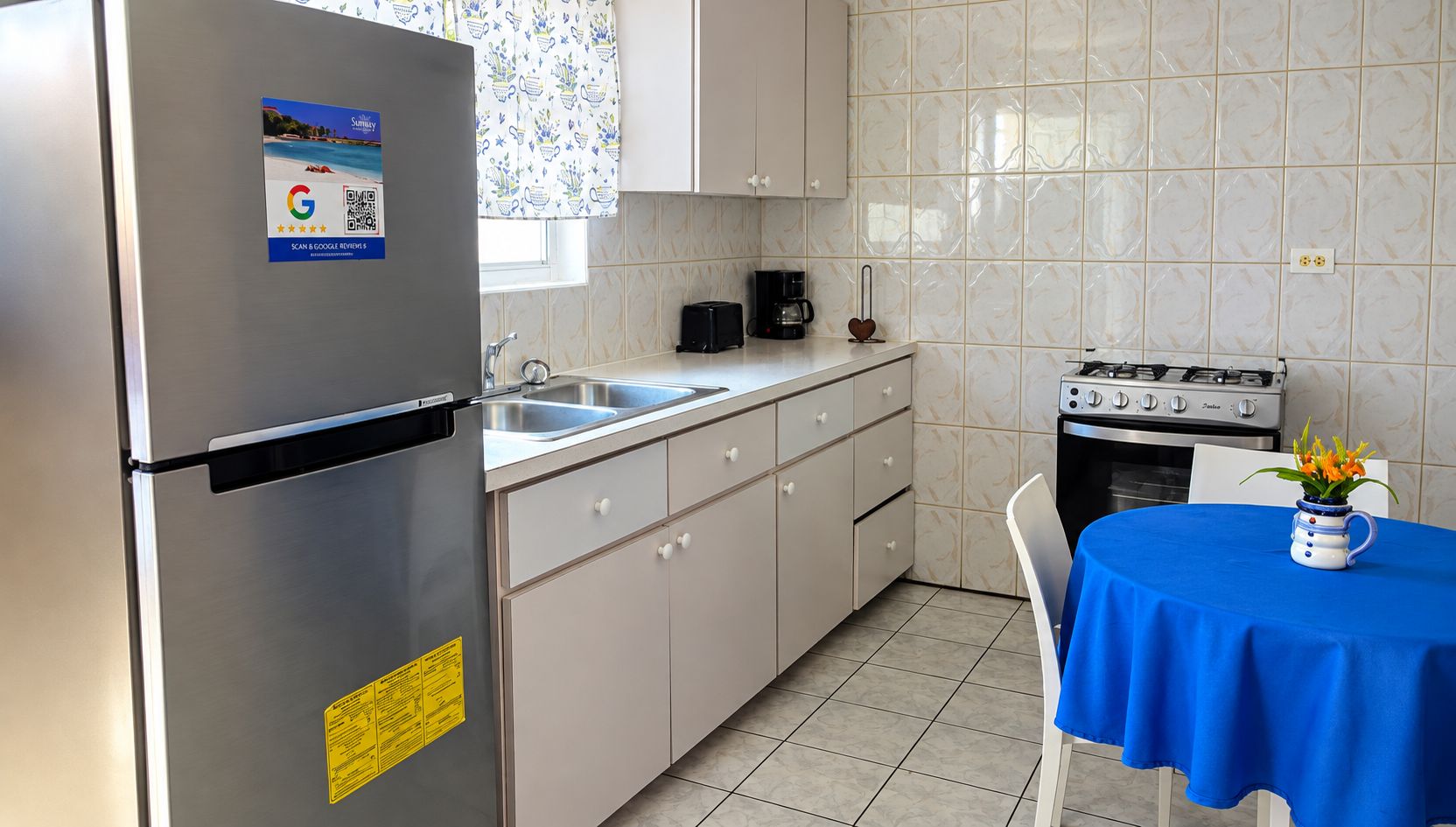 Cozinha compacta com geladeira de aço inoxidável, cafeteira, fogão e mesa com toalha de mesa azul.
