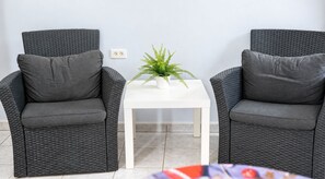 Duas cadeiras de vime cinzentas com almofadas, uma mesa branca com uma planta e um chão de azulejos.