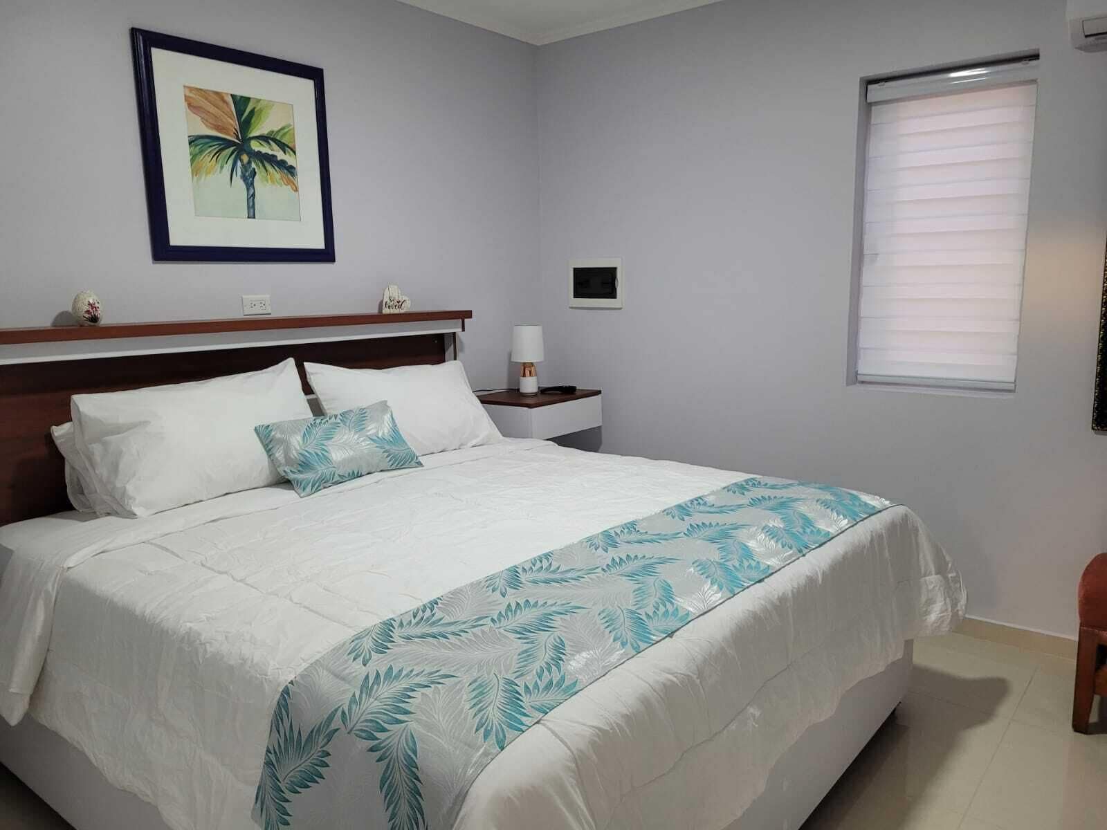 A neatly made bed with a white and teal leaf-patterned bedspread, a framed tropical artwork on the wall, a bedside table with a lamp, and a window with blinds.