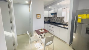 A compact kitchen with white cabinets, a black countertop, a stainless steel refrigerator, and a small dining table with chairs. - Sunway Aruba Aparments (Santa Cruz)