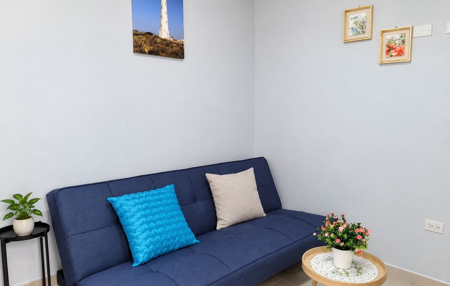 Um sofá azul com duas almofadas, uma mesinha redonda com uma planta em vaso e um quadro na parede.