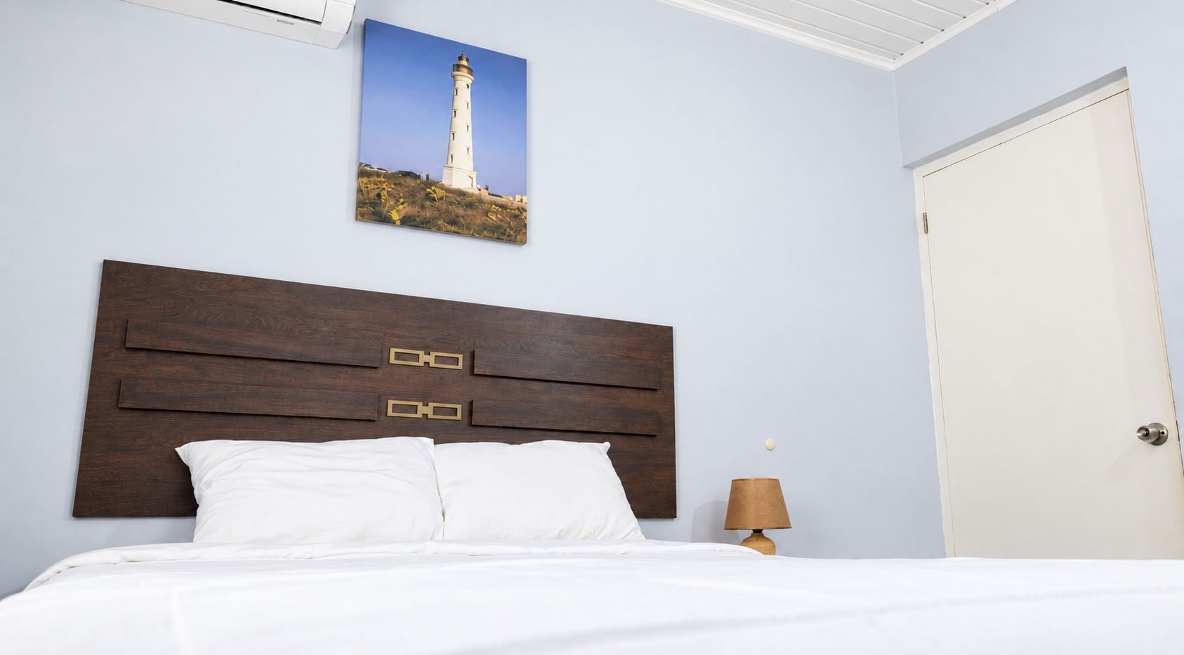 Une chambre à coucher avec une tête de lit en bois, un lit recouvert de linge de lit blanc, une table de chevet avec une lampe et une porte.