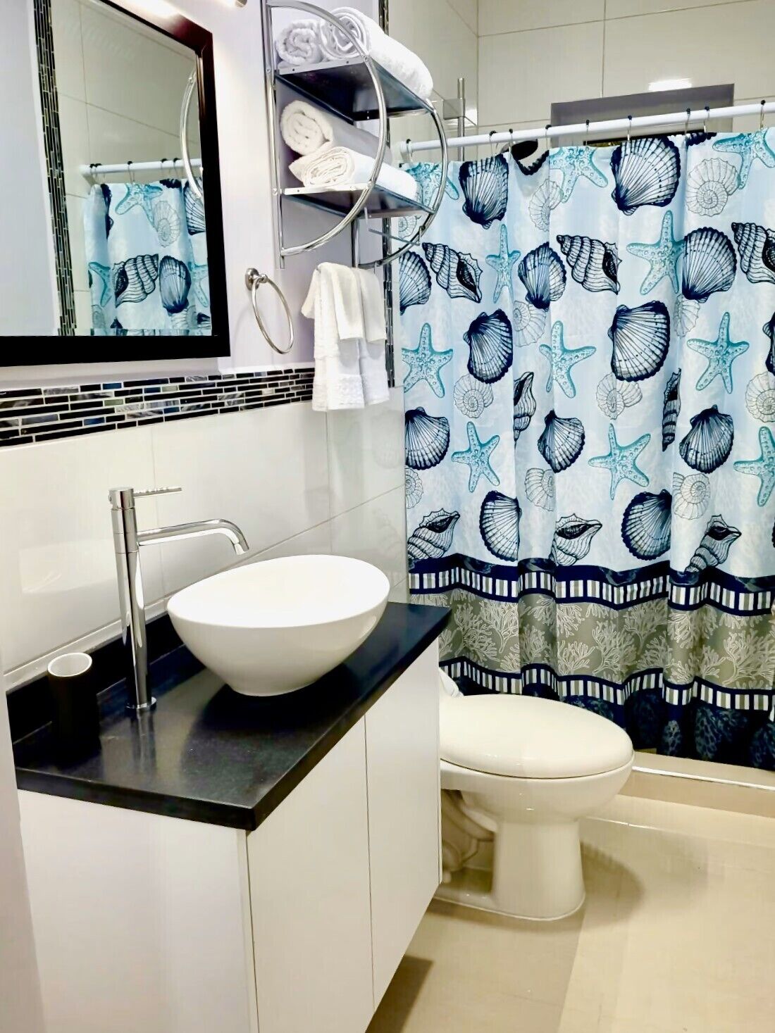Un baño con lavamanos blanco, un espejo, un inodoro y una cortina de ducha con estampado de conchas marinas.
