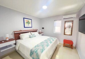 A bedroom with a bed, nightstands, a red bench, a wall-mounted TV, a window, and a framed picture. - Sunway Aruba Aparments (Santa Cruz)