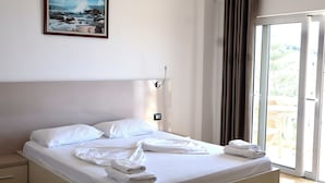 A neatly made bed with white linens and towels, a painting on the wall, and a view through a sliding glass door. - Mucobega Hotel  (Sarande)