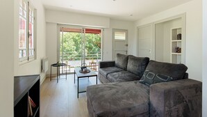 A living room with a grey sofa, a small table, and a radiator. There is a door and a window with blinds. - Appartement Prunelle - Welkeys (Soorts-Hossegor)