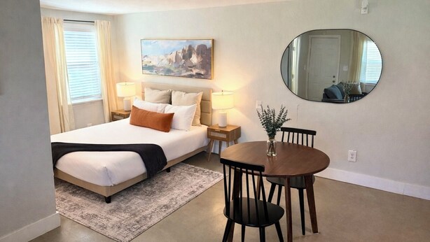 Une chambre d’hôtel avec un lit, des tables de chevet, une table ronde avec des chaises, un miroir rond et un tableau au mur.