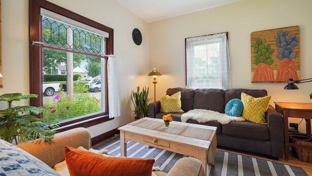 A living room with a brown sofa, a coffee table, and a window with a view of a car and greenery outside. - Charming 2-bedroom house in Viroqua with AC (Viroqua)