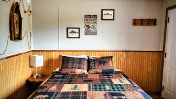 A bedroom with a wooden headboard, a quilted bedspread, a bedside lamp, and wall decorations including framed fish and fishing-related signs.