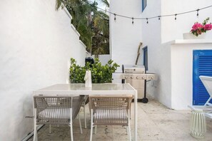 A white outdoor dining area with a table and two wicker chairs, a grill, and a blue door in the background. - Ocean Park 1BD-1BA-Pool Walktobeach -V (San Juan)