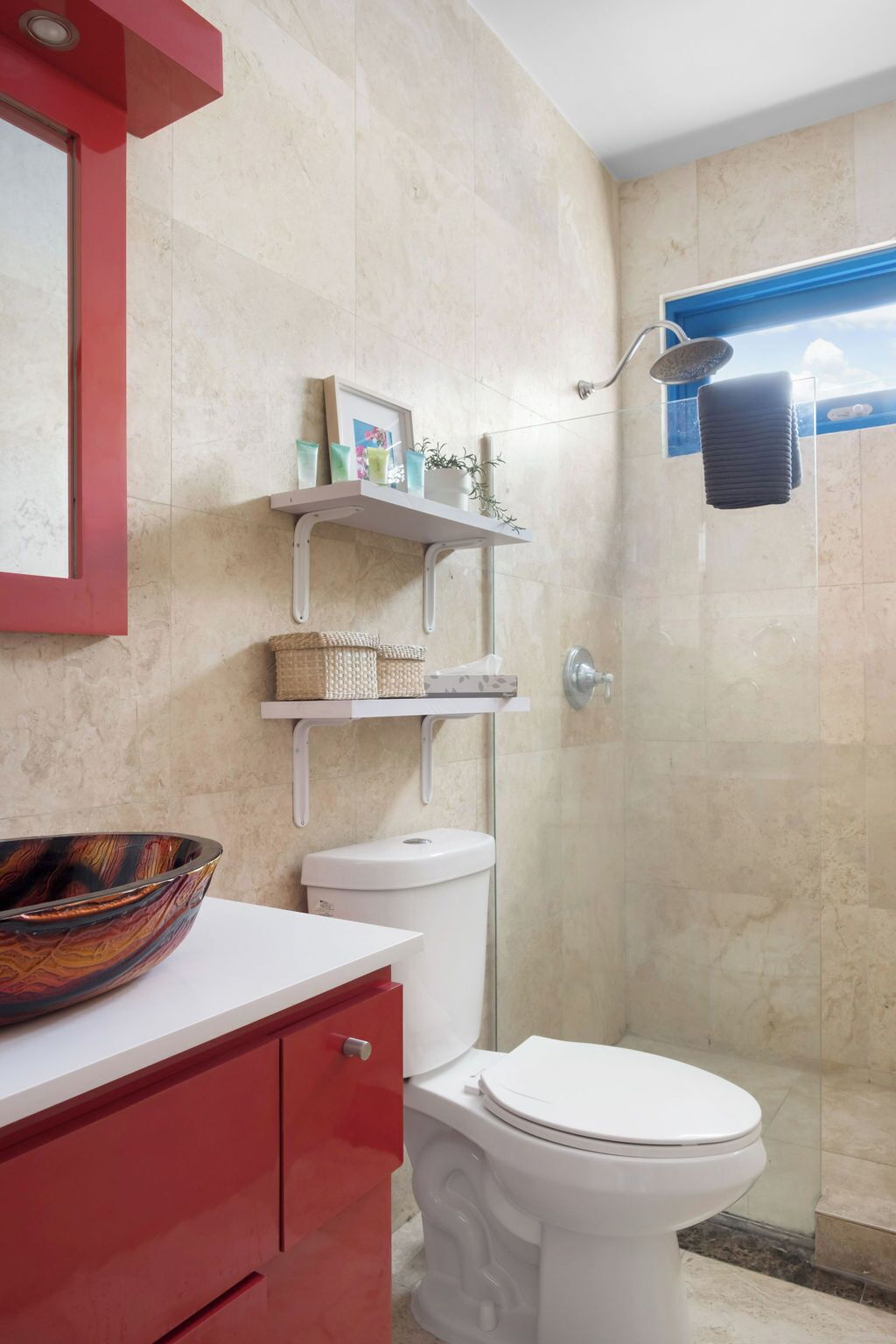 Bany amb vàter blanc, un armari vermell, una cabina de dutxa de vidre i una prestatgeria amb productes de tocador.