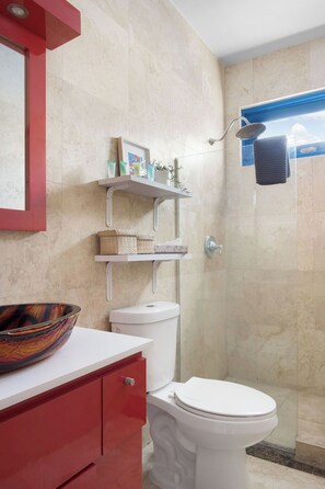 A bathroom with a white toilet, a red cabinet, a glass shower enclosure, and a shelf with toiletries. - Ocean Park 1BD-1BA-Pool Walktobeach -V (San Juan)
