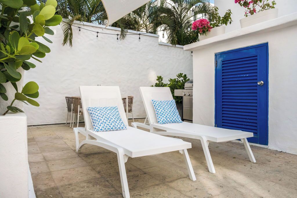 Dos sillones blancos con cojines estampados, una puerta azul y una pared blanca con plantas y luces de adorno.