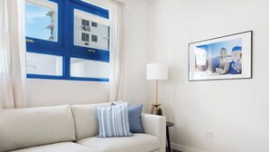A modern living room with a beige sofa, a framed picture of a coastal scene, a lamp, and a window with blue shutters. - Ocean Park 1BD-1BA-Pool Walktobeach -V (San Juan)