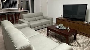 Uma sala de estar moderna com um amplo sofá em formato de L, uma mesa de centro de madeira, uma televisão de tela plana e um lustre.